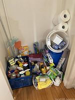 Wide view of cleaning supplies lot showing blue crate with sprays, sponges, cloths, bucket, mop, and trash bags piled against wall