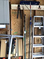 Photo showing a 6-foot aluminum step ladder with four steps, garden shovel with blue handle grip, large metal framing square, and two black-handled loppers hung on a wood wall.