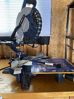 Overall side view of the blue and gray sliding compound miter saw on a wooden surface with some paper on the stand.