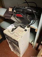 Craftsman belt sander sitting on shelf with extra sandpaper rolls and user manual on top of original box below.