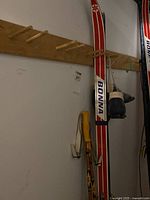 Full view of Bonna cross country skis, a pair of ski poles with yellow handles, and a pair of black and white cross country ski boots hanging on wooden pegs