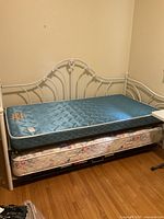 Full view of white metal day bed frame with blue single mattress and floral trundle mattress underneath