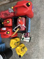 Top view of collection showing multiple red plastic fuel cans with yellow caps, siphon pump with flexible hose, two yellow Pennzoil motor oil jugs, Sta-Bil and STP fuel stabilizer bottles on concrete floor.