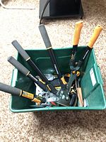 Green plastic bucket containing Fiskars long-handled loppers, hand pruners and other garden clippers with black and orange handles.