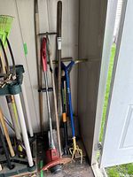Wide view of garden tools arranged in a corner, including long-handled shovels, rakes, electric weedeater, cultivators, with a partially open door showing outdoor grass.