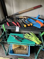 Photo showing long-handled loppers, several small hand tools, blue dustpan, and garden stool with gloves on top.