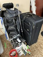 Black golf bag standing with visible golf clubs and next to black luggage.