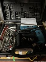 Photo of Makita hammer drill in black carrying case with multiple drill bits and accessories inside the case compartments, and bagged bits on the side.