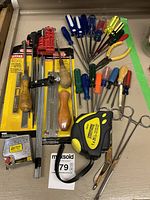 Wide view of assorted hand tools including screwdrivers with plastic handles in various colors, pliers, needle files, tape measure, and other small hand tools on a beige surface.