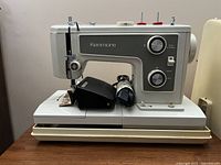 Side profile showing the Kenmore brand label alongside foot pedal and power cord on base of machine with beige case below