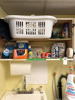 Photo of laundry shelf with laundry basket on top and laundry and cleaning products arranged on shelf including detergents, bleach powder, fabric softener and spray bottles.