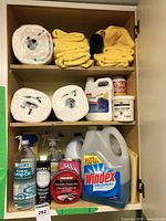Full view of cupboard with cleaning supplies including towels, paper towels, Windex, lamp oil, and gel cleaner.