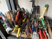 Wide shot of various hand tools arranged on a shelf including pliers, wire clippers, scrapers, and a hammer with colored handles.