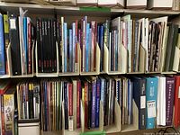 Full shelf view showing a large collection of books and magazines related to quilting, sewing, and cross stitch projects, organized standing vertically with spine titles visible.