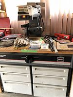 Full view of radial arm saw on workbench with accessories including saw blades, gloves, manual, hand tools, and containers arranged on the surface.