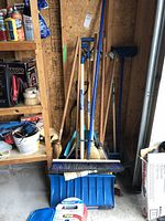 Photo showing a collection of snow shovels with blue plastic scoop blades and multiple brooms with wooden handles in a garage corner.