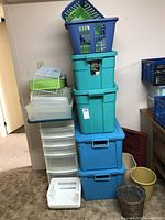 Stack of different colored plastic storage totes, white and green drawer organizers, and small plastic baskets on carpeted floor against wall.