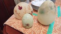 Three decorative egg-shaped candles of different sizes on a table with stacked dishes behind them.