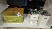 Display of vintage breadbox and three white enamel canisters with metal lids on shelf, showing overall condition and details including labels on canisters and decorative text on breadbox lid.