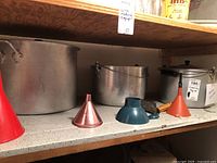 View of multiple funnels in red, pink, blue, and orange plastic, one metal funnel, two large aluminum pots, and a metal pail on a shelf.