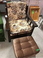 Wooden frame chair with floral upholstery and brown buttoned ottoman placed in a basement room with other furniture.