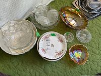 Multiple China and glass dishes including Royal Albert cake plate, Carnival glass bowls and clear pressed glass serving dishes laid out on green surface.