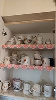 Shelves with various cups, mugs, creamers, coffee pot, and other china and pottery items arranged in a cabinet with lace shelf liners.