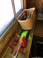 Overview of three large red pipe clamps and a woven basket with various hand tools on a wooden bench by a window in a barn or shed setting.