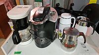 Photo showing two coffee makers, a Keurig single serve and a white drip coffee maker, next to two insulated flasks and a stainless steel coffee pot with floral design.