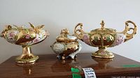 Three-piece decorative porcelain set with a gold-tone finish, pink floral designs, and ornamental handles. The compote is on the left, the two covered dishes are in the center and right.