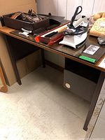 Wooden desk table with glass top, shown with items on top. Desk has dark brown finish and a drawer or compartment under the glass top. Visible wear including scratches and chips.