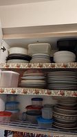 Multiple shelves showing stacked white and light-colored stoneware plates, bowls, and saucers alongside various plastic food storage containers with lids.