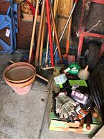 Full lot showing clay pots, various hand tools with wooden handles, green garden sprayer, gloves, bottles of chemicals on cardboard box beside a wheelbarrow.