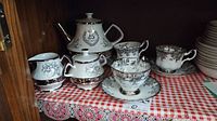 Gibson china teapot, sugar bowl, creamer jug, and Royal Albert teacups and saucers displayed on shelf with red and white checkered cloth