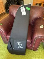Full view of the black Gibson guitar case standing upright on a red leather armchair with visible Gibson logo on the front pocket.