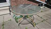 Round glass top metal table with rusty beige metal frame and legs, placed outdoors on stone tiles near brick wall and garden hose.