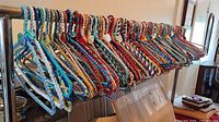 Photo showing a large number of artisan craft wire hangers covered with multicolored woven fabric hanging on a rod, with two plastic blue hangers visible on the left.