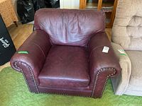 Front view of dark reddish-brown leather armchair with nail head trim on rolled arms, showing leather wear and creasing.