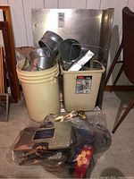 View of the two plastic buckets, one cream and one beige, filled with multiple metal baking-related pans and tins. Also includes a metal tea strainer.