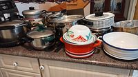 Wide view of various pots and pans including metal and enamel items, lids, and kitchen containers on countertop.