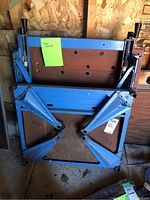 Blue metal sawhorse folded up with visible brown support panel and adjustable crossbars, showing signs of wear and some rust spots.