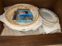 Photo of ceramic plates and bowls stacked with a wicker basket containing boxed cutlery sets on top