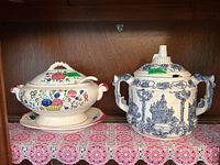 Two soup tureens side by side on a wooden shelf with a pink lace doily underneath. Left tureen has colorful floral design with ladle and saucer. Right tureen is blue and white with architectural patterns and double handles.