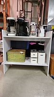Storage table with kitchen appliances and containers arranged on two shelves. The table is white with visible scratches. Kitchen appliances include coffee machines and electric kettle on top. Bottom shelf has vintage bread box and labeled containers.