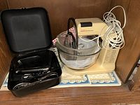 Sunbeam Mixmaster cream-colored vintage stand mixer with glass bowl, two metal beaters, and Black & Decker black hand mixer stored in black case with power cord and accessories, all set on a kitchen cabinet shelf.