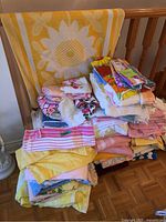Stack of towels on a wooden chair. Large yellow towel with white sunflower design behind the pile. Various pink, floral, striped, and yellow towels stacked.