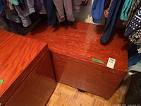 Wooden trunk placed in a closet next to hanging clothes, showing top and front sides with reddish-brown polished wood grain.