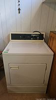 Front and side view of a vintage white Kenmore dryer in basement with control knobs on top panel.