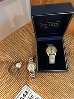 Three wristwatches arranged on a wicker surface, including one Seiko automatic watch inside its original Seiko box, a Basa watch with gold-tone case, and a Timex watch with gold-tone case.