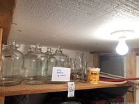 Wide shelf view showing five large clear glass wine jugs, plastic hoses coiled next to a yellow bucket, and a funnel.